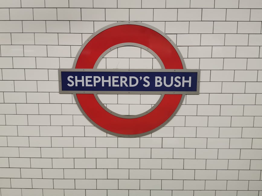 A close-up view of the Shepherd's Bush London Underground station sign mounted on a white tiled wall. The sign features a circular red border with a horizontal blue bar across the middle, displaying the station name 'SHEPHERD'S BUSH' in white uppercase letters. This image is taken inside an underground station corridor, with the sign positioned centrally against the wall of uniformly arranged rectangular tiles. The signage indicates the station's location within the tube network and is relevant to the area served by Shepherds Bush Man and Van, which offers house removals and furniture transport services. The clean, well-lit environment emphasizes the station's branding and provides context for home relocation and transport logistics.