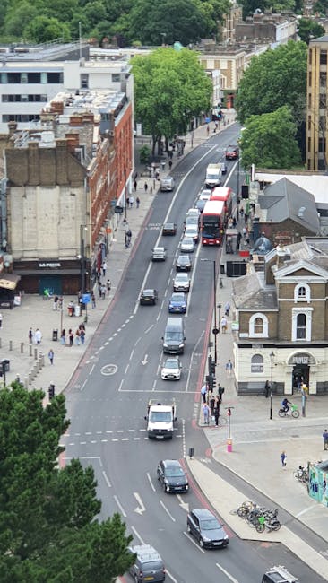 An aerial view of a busy street in Shepherds Bush, showing vehicles including cars, vans, and a double-decker bus moving along the road. On the pavement, pedestrians are walking near storefronts and residential buildings, with some people waiting at pedestrian crossings. The scene features a mix of older brick and modern buildings, with green trees lining the street, and a small white church-style building at the corner. In the background, there are more trees and residential rooftops. This image captures the typical urban environment where house removals and furniture transport activities by Shepherds Bush Man and Van take place, emphasizing the importance of careful logistics during home relocation or moving processes that involve loading or unloading near the street.